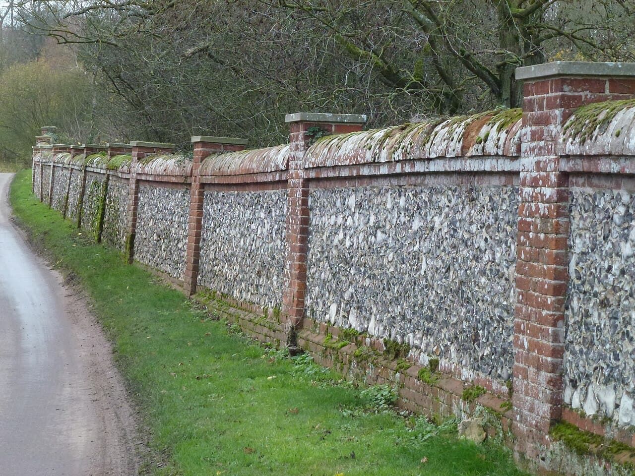 A knapped-flint boundary wall with brick piers running along a lane.